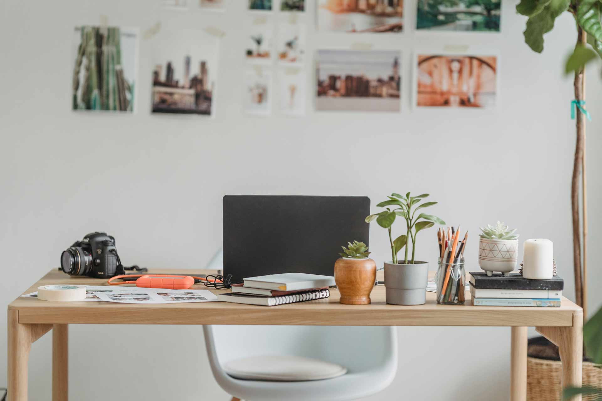 bureau de travail, avec un ordinateur portable, un appareil photo, des carnets & livres. Ainsi que diverses plantes. En fond nous pouvons voir un mur où sont accrochés des visuels & illustrations.