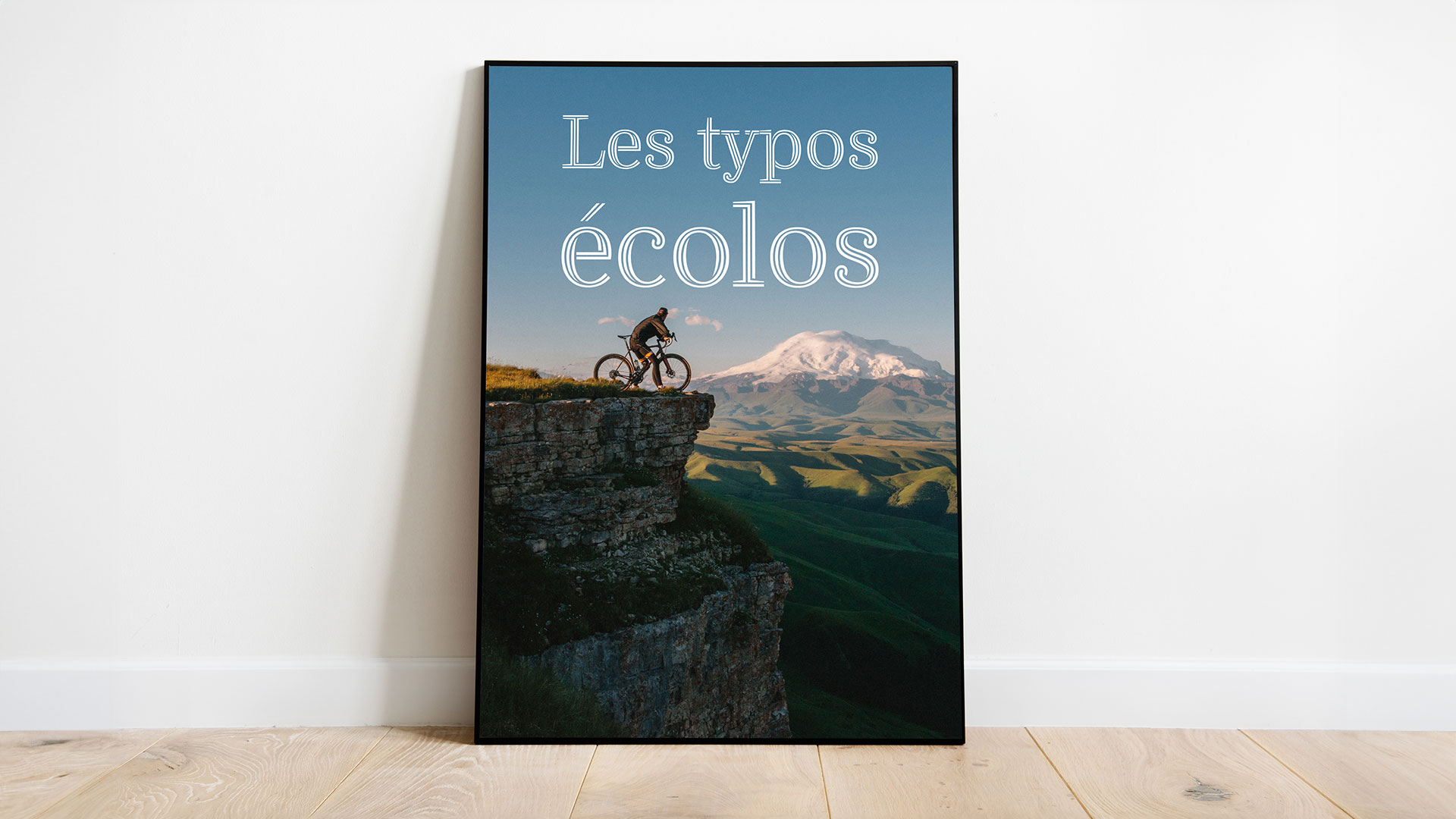 Cadre posée sur le sol et contre un mur. Nous voyons dans le cadre un vélo et son cycliste au bord d'une falaise. La mention "les typos écolos" est inscrite sur le ciel au dessus du cycliste